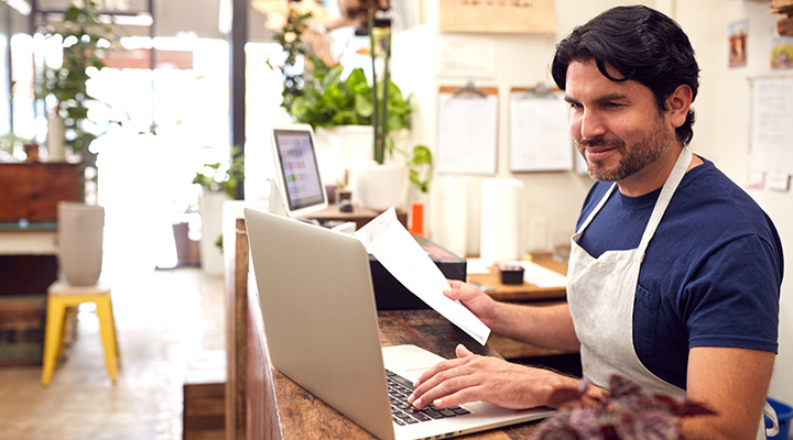Man in front of laptop and wearing an apron while holding paper document